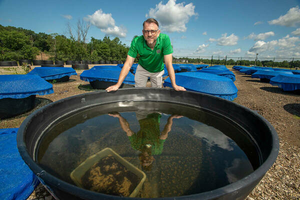 Jason Rohr stands in front of large black cylinder filled with water; his reflection shines on the water