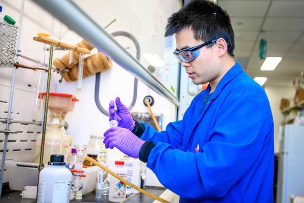 Researcher in blue lab coat and goggles holds vial in lab