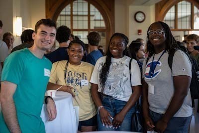 Four Notre Dame students smile for a photo in a building with arched doorways.  The student on the left wears a teal shirt, the two center students wear gold and white shirts, and the student on the right wears a grey shirt with a Champion logo.