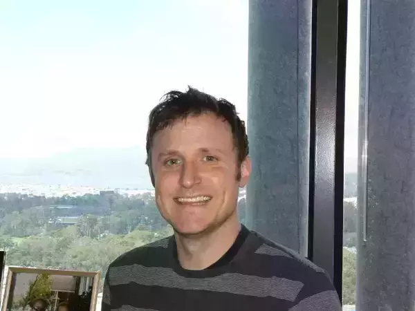 A man with short dark hair and a wide smile, wearing a dark grey striped shirt, stands before a window overlooking a bright city, green hills, and a bay.