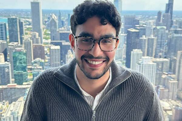 Mohamed El Gazzah, smiling, in a grey ribbed sweater and glasses overlooks the Chicago skyline from a high-rise.
