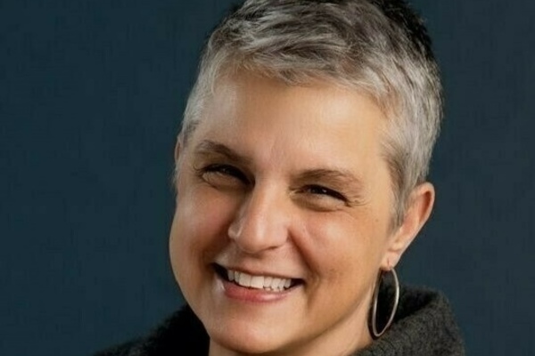 Jennifer Tank smiles, with short gray hair, wearing a dark gray sweater and a silver hoop earring, against a dark blue background.