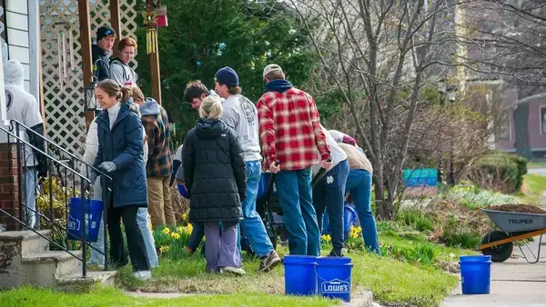Students do spring yard work. One woman smiles carrying a blue bucket, others fill a wheelbarrow with mulch. Daffodils bloom.