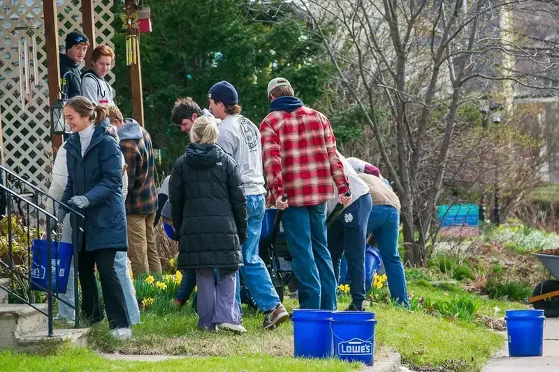 Students do spring yard work. One woman smiles carrying a blue bucket, others fill a wheelbarrow with mulch. Daffodils bloom.