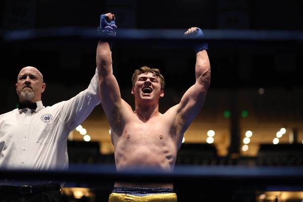 Boxer Jerry Rullo wears gold shorts & blue wraps and raises arms triumphantly as a referee in a Notre Dame Boxing shirt declares victory. Heavyweight division in the 2023 Bengal Bouts.