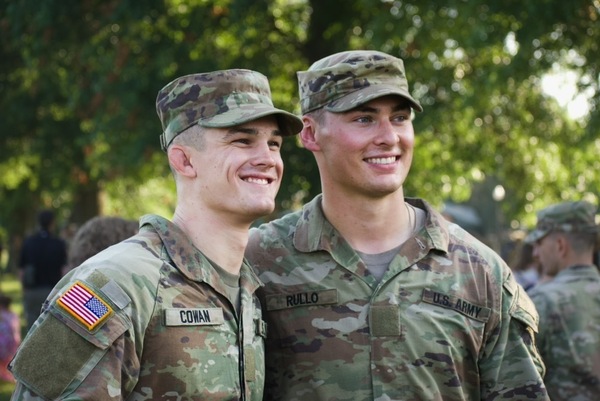 Two smiling men, Cowan and Rullo, in US Army camouflage uniforms and patrol caps stand outdoors. Cowan has a US flag patch. Notre Dame ROTC