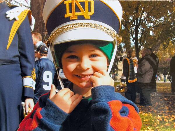 Jerry Rullo as a child in a Notre Dame band hat over green beanie, blue/orange fleece, stands by a band member.
