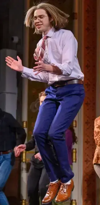 Adam Albritton with shoulder-length blonde hair and glasses jumps mid-air, smiling, wearing a white shirt, pink tie, blue pants, and brown shoes.
