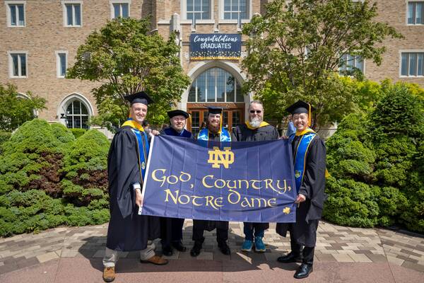 Five people holding a flag that reads 'God, Country, Notre Dame' while looking at the camera.