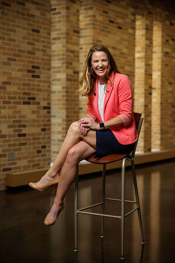 Kate Biberdorf, with brown hair, in coral blazer, white top, navy skirt, sits on a stool with hands clasped.