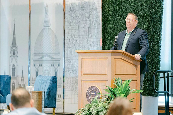 A man in a navy blazer and green shirt stands at a wooden podium with the University of Notre Dame seal. He speaks at a microphone with a backdrop featuring line art of the Basilica of the Sacred Heart and a historical image.