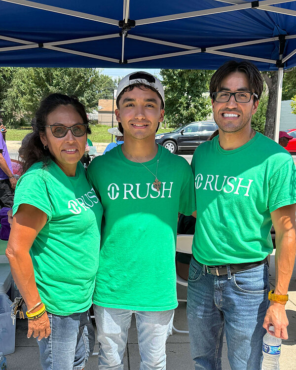 Three smiling people stand under a blue tent, all wearing green "RUSH" t-shirts.
